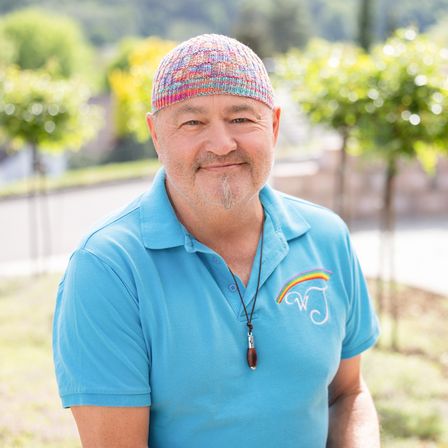 Ein Mann mit einem blauen Poloshirt mit Regenbogenlogo und einer bunten gestrickten Mütze lächelt in einer Außenumgebung.