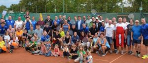 Eine Gruppe von Menschen posiert für ein Foto auf einem Tennisplatz, hält Tennisschläger und einige tragen Sportkleidung. Bäume und ein grüner Zaun sind im Hintergrund.