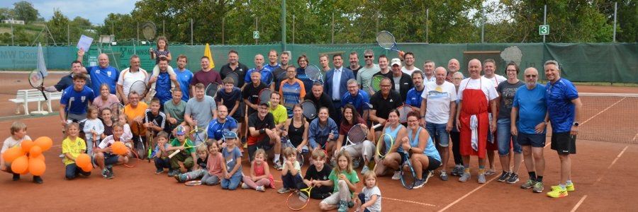 Eine Gruppe von Menschen posiert für ein Foto auf einem Tennisplatz, hält Tennisschläger und einige tragen Sportkleidung. Bäume und ein grüner Zaun sind im Hintergrund.