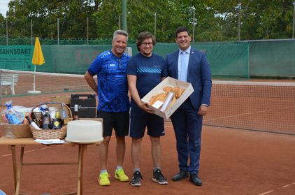 Drei Männer stehen auf einem Tennisplatz, einer hält eine Brotbox, ein anderer eine Flasche und ein Dritter lächelt. Ein Tisch mit einem Korb und einem Kuchen steht in der Nähe. Im Hintergrund befindet sich ein grüner Zaun mit Bäumen.