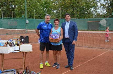 Drei Personen stehen auf einem Tennisplatz. Der Mann links trägt ein blaues Shirt und gelbe Turnschuhe. Die Frau in der Mitte hält einen Korb. Der Mann rechts trägt einen Anzug und eine Krawatte. Sie alle lächeln. Hinter ihnen steht ein Tennisnetz. Links befindet sich ein Tisch mit einem weißen Tuch und Flaschen. Bäume stehen hinter dem Zaun.