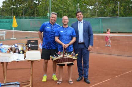 Drei Männer auf einem Tennisplatz, einer hält einen Korb mit Flaschen. Ein junges Mädchen steht im Hintergrund.