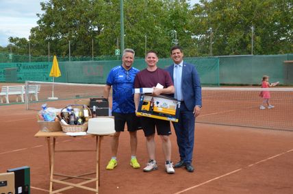 Drei Männer stehen lächelnd auf einem Tennisplatz. Einer hält eine Schachtel mit einem gelben Spielzeuglastwagen. Ein anderer hält ein Papier. Verschiedene Gegenstände stehen auf einem Tisch in der Nähe, und ein gelber Schirm steht hinter ihnen. Bäume und ein grüner Zaun sind im Hintergrund.