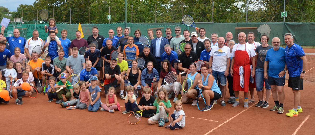 Eine große Gruppe von Menschen, einschließlich Kindern, posiert für ein Foto auf einem Tennisplatz. Sie halten Tennisschläger und einige sitzen, während andere stehen. Bäume und ein Zaun sind im Hintergrund zu sehen.