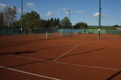 Ein leerer Tennisplatz mit rotem Lehmboden und weißen Linien. Im Hintergrund befindet sich ein Zaun mit Werbetafeln. Bäume und Straßenlaternen sind in der Ferne zu sehen.