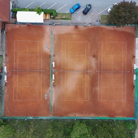 Luftaufnahme eines Tennisplatzes mit drei Plätzen. Die Plätze sind leer und haben eine braune Oberfläche. Bänke und Bäume sind um den Platz herum sichtbar.