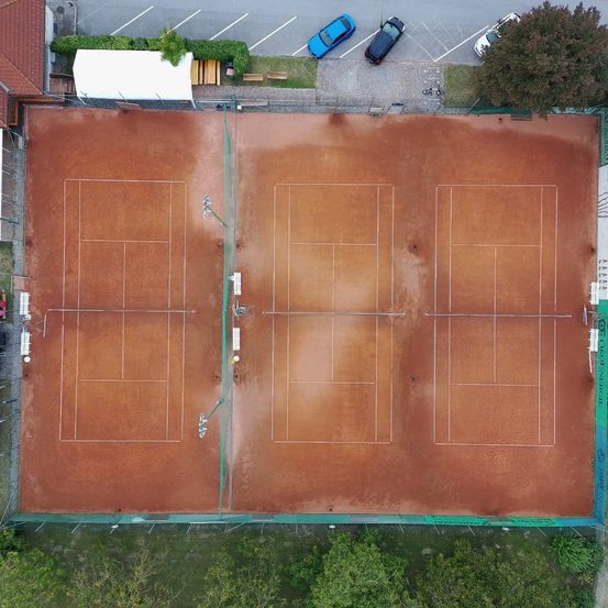 Luftaufnahme eines Tennisplatzes mit drei Plätzen. Die Plätze sind leer und haben eine braune Oberfläche. Bänke und Bäume sind um den Platz herum sichtbar.