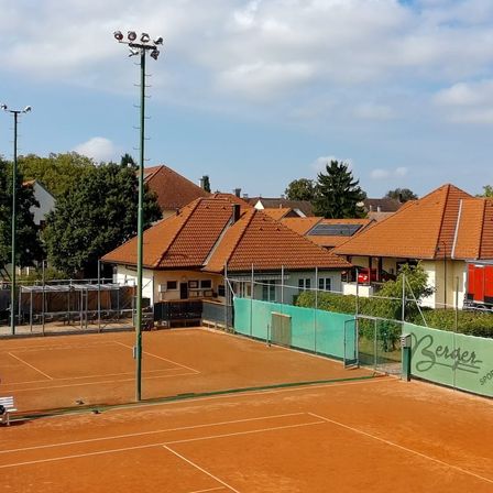 Ein leerer Tennisplatz mit rotem Belag ist von einem grünen Zaun umgeben. Der Platz wird von einem hohen Laternenmast beleuchtet und ist von einem Metallzaun eingeschlossen. Dahinter befinden sich Gebäude mit braunen Dächern.