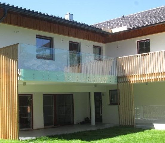 Ein modernes Haus mit einem Glasbalkon und Holzgeländern. Das Haus hat ein Ziegeldach und große Fenster. Es ist von einem Rasen umgeben.