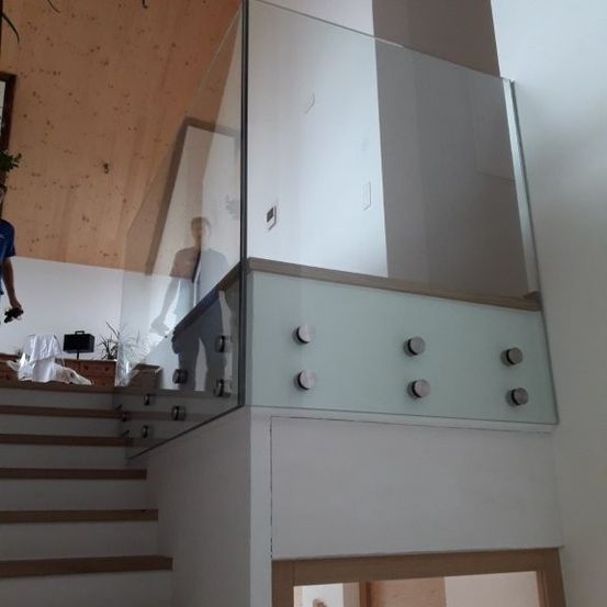 Ein Glasgeländer mit Metallpfosten ist an der Treppe installiert. Eine Person steht in der Nähe des Geländers, und ein Holzrahmen befindet sich an der Wand. Ein Topf mit einer Pflanze ist auf dem Holzschrank platziert. Eine Person steht auf der linken Seite.