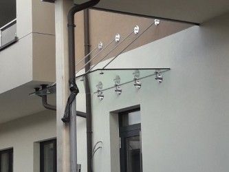 Ein Gebäude mit einem Glasvordach, das von Metallstangen gestützt wird, mit einem Rohr auf der linken Seite und einem Balkon darüber. Das Vordach hat mehrere Lichter und ist an einer weißen Wand befestigt.