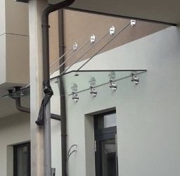 Ein Gebäude mit einem Glasvordach, das von Metallstangen gestützt wird, mit einem Rohr auf der linken Seite und einem Balkon darüber. Das Vordach hat mehrere Lichter und ist an einer weißen Wand befestigt.