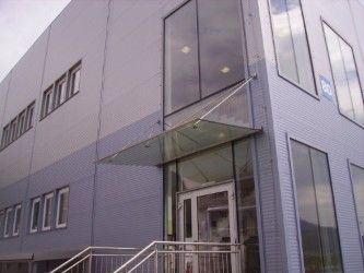 Ein graues Gebäude mit Glasfenstern und einem Glasvordach über dem Eingang. Das Gebäude hat mehrere Stockwerke und mehrere Fenster. Der Eingang ist mit einer weißen Tür und einem Glasvordach ausgestattet.