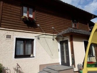 Ein Holzhaus mit einer weißen Wand und einer schwarzen Tür. Ein gelbes Fahrzeug ist vor dem Haus geparkt. Es gibt ein Fenster mit einem Blumentopf und einem Fenstergitter.