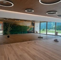 Ein leerer Raum mit großen Fenstern, einem Holzboden und einem Wandbild eines Baumes. Es gibt runde Deckenleuchten und Reflexionen in der Glasscheibe.