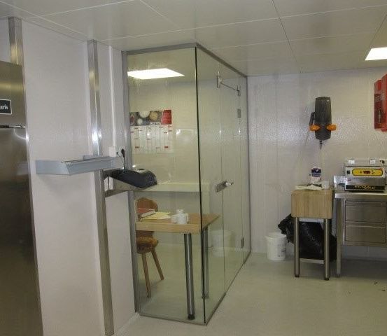 Ein kleiner Küchenbereich mit einer Glastrennwand, einem Schreibtisch und einem Stuhl. Der Raum umfasst einen Kühlschrank, einen Tisch und verschiedene Geräte.