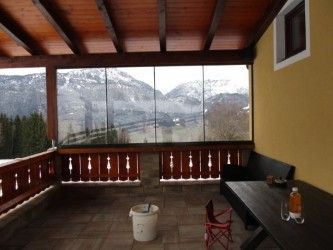 Ein Balkon mit Blick auf verschneite Berge. Eine Holzbank und ein roter Stuhl stehen neben einem Tisch mit einer Flasche darauf. Der Balkon hat einen gefliesten Boden und ein hölzerne Geländer.