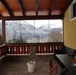Ein Balkon mit Blick auf verschneite Berge. Eine Holzbank und ein roter Stuhl stehen neben einem Tisch mit einer Flasche darauf. Der Balkon hat einen gefliesten Boden und ein hölzerne Geländer.