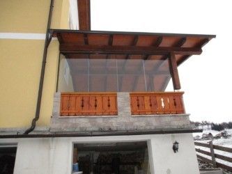 Ein Balkon mit einem braunen Holzdach und Geländern ist an einem gelben Gebäude befestigt. Der Balkon hat Glasscheiben, und eine Lampe ist an der Wand montiert.