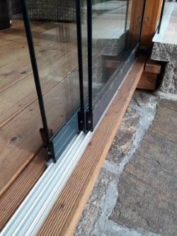Ein Nahaufnahme einer Glastür mit schwarzen Rahmen und hölzernen Schwellen. Die Tür ist leicht geöffnet und zeigt einen Holzboden und eine Steinstufe.