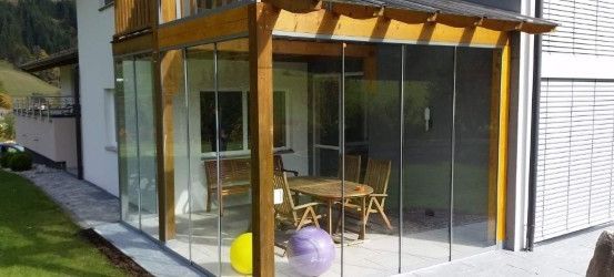 Ein modernes Haus mit einer Glasterrasse und Holzmöbeln. Ein Tisch mit Stühlen steht in der Nähe der Glaswand. Zwei bunte Bälle liegen auf dem Boden.
