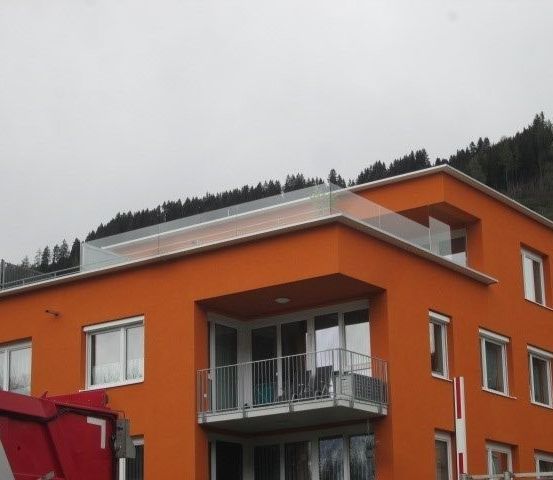 Ein orangefarbenes Gebäude mit Glasgeländer auf dem Dach und Balkon. Das Gebäude hat mehrere Fenster und einen Balkon mit Stühlen. Ein roter Maschinenwagen ist in der Nähe geparkt.