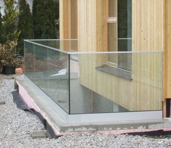 Ein Hausaußenbereich mit einer Holzwand und Glasgeländern ist zu sehen. Bäume und Pflanzen sind im Hintergrund sichtbar.