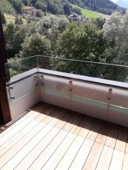 Ein Balkon mit einer Glasbrüstung bietet einen Blick auf ein üppig grünes Tal mit Bergen in der Ferne. Der Holzboden und die Bäume sind deutlich zu erkennen.