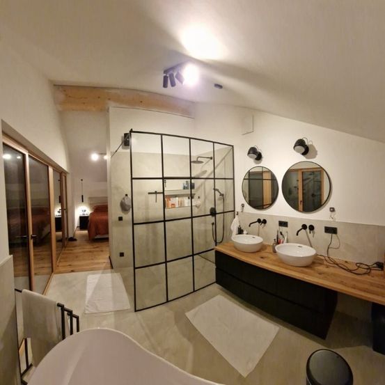 Ein modernes Badezimmer mit einer Glasdusche, zwei weißen Waschbecken und zwei runden Spiegeln. Der Raum verfügt auch über eine Holztheke und eine große weiße Badewanne. Es gibt weiße Handtücher und Teppiche auf dem Boden.