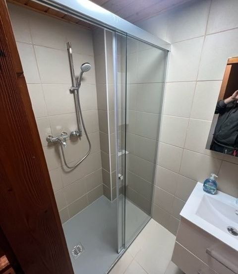 Ein Badezimmer mit einer Glasdusche, gefliesten Wänden und einem Waschbecken mit einer Flasche Handseife. Es gibt eine Holztür und einen Spiegel, der eine Person reflektiert.