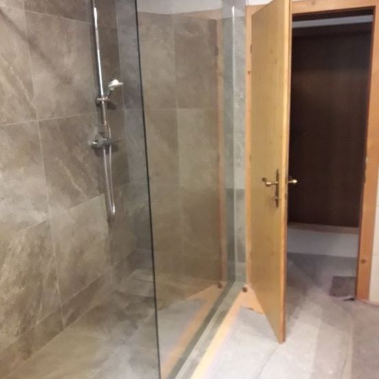 Das Bild zeigt ein Badezimmer mit einer Glasdusche. Die Wände und der Boden sind mit einem hellgrauen Muster gefliest. Die Duschbrause ist an der Wand befestigt, und das Wasser ist abgestellt.