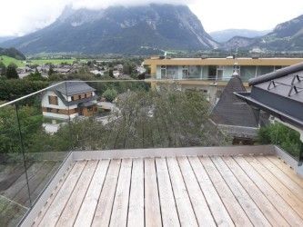 Ein Balkon mit Holzboden und Glasgeländern, mit Blick auf eine Stadt mit Bergen in der Ferne. Es sind zwei Häuser zu sehen, eines mit braunem Dach und das andere mit gelber Fassade.