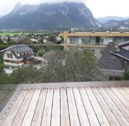 Ein Balkon mit Holzboden und Glasgeländern, mit Blick auf eine Stadt mit Bergen in der Ferne. Es sind zwei Häuser zu sehen, eines mit braunem Dach und das andere mit gelber Fassade.