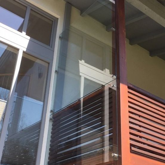 Ein Balkon mit Glasgeländern und einem hölzernen Deck, angeschlossen an ein modernes Gebäude mit großen Glasfenstern und einem beigefarbenen Äußeren.