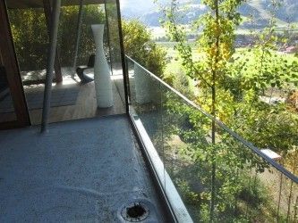 Ein Balkon mit einer Glasbrüstung bietet einen Blick auf eine ruhige Landschaft mit Bäumen und einer entfernten Aussicht auf Berge. Eine weiße Vase und ein Stuhl stehen auf dem Balkon.