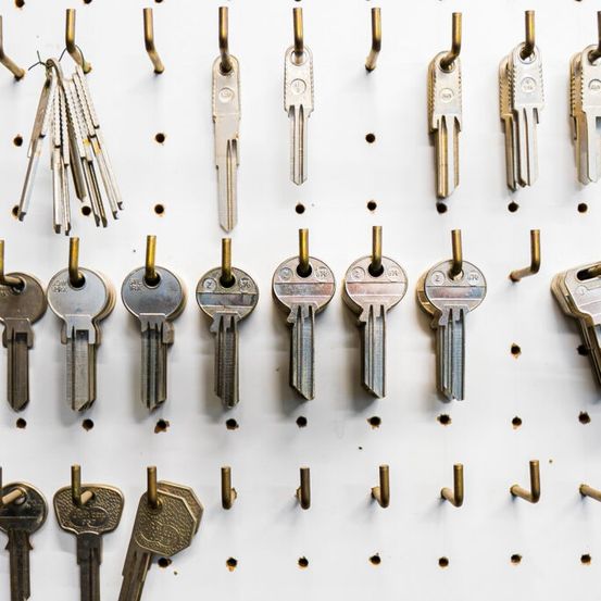 Ein weißes Pegboard zeigt eine Sammlung verschiedener Schlüssel, die jeweils mit einem goldenen Haken befestigt sind. Die Schlüssel sind in Reihen angeordnet und präsentieren eine Vielzahl von Stilen und Größen.