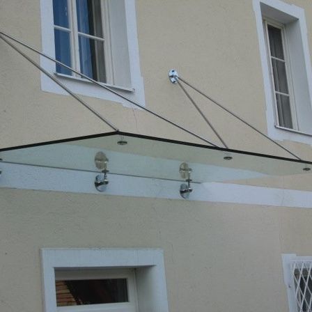 Ein Glasvordach ist an der Wand eines Gebäudes mit zwei Fenstern befestigt. Das Vordach hat einen Metallrahmen.