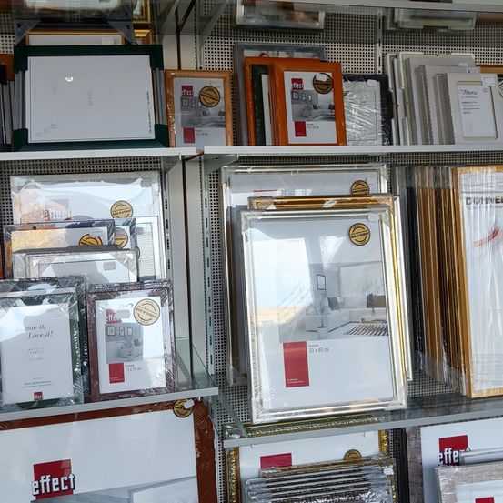 Ein Ladendisplay mit Regalen, die zahlreiche Bilderrahmen enthalten, sowohl leere als auch einige mit Beispielbildern. Die Rahmen variieren in Farbe und Stil, wobei einige goldene Akzente aufweisen. Die Marke des Geschäfts ist sichtbar, und die Regale sind gut organisiert.
