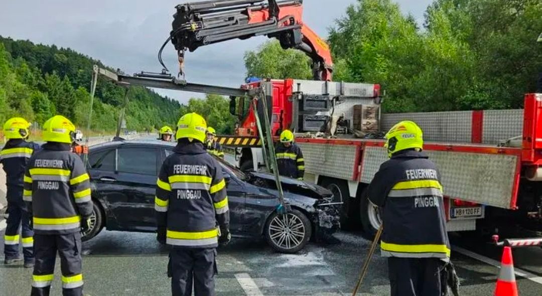 Feuerwehrleute heben ein demoliertes Auto mit einem Kran. Zwei Feuerwehrleute stehen in der Nähe des Autos. Der Kran ist rot.