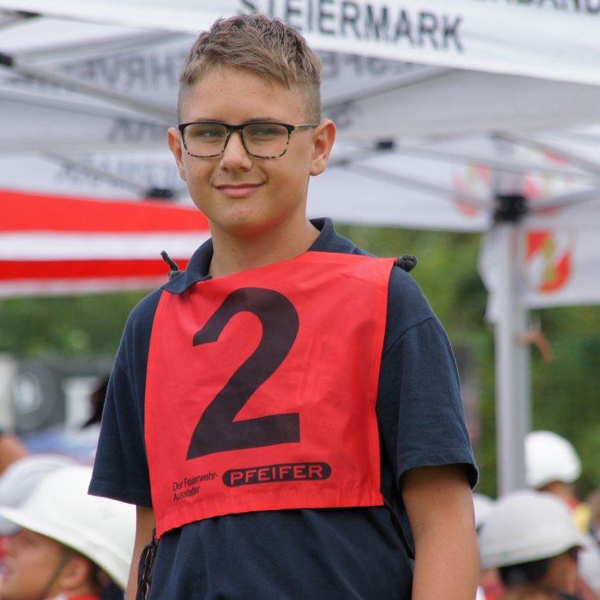 Ein junger Junge mit Brille steht draußen und trägt ein rotes Weste mit der Nummer 2 und ein blaues Polohemd. Im Hintergrund ist ein weißes Zelt mit dem Wort Steiermark zu sehen.