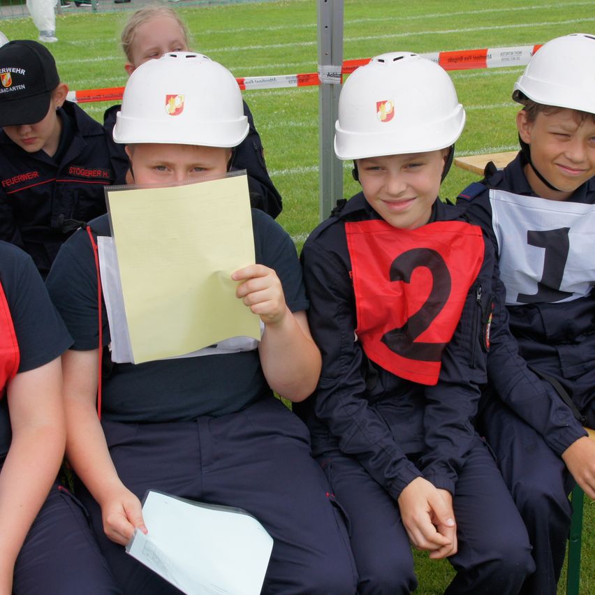 Vier Kinder in blauen Uniformen und weißen Helmen sitzen auf einer Bank, eines hält ein gelbes Papier und ein anderes trägt eine rote Weste mit der Nummer 2. Sie scheinen an einem Wettbewerb teilzunehmen.