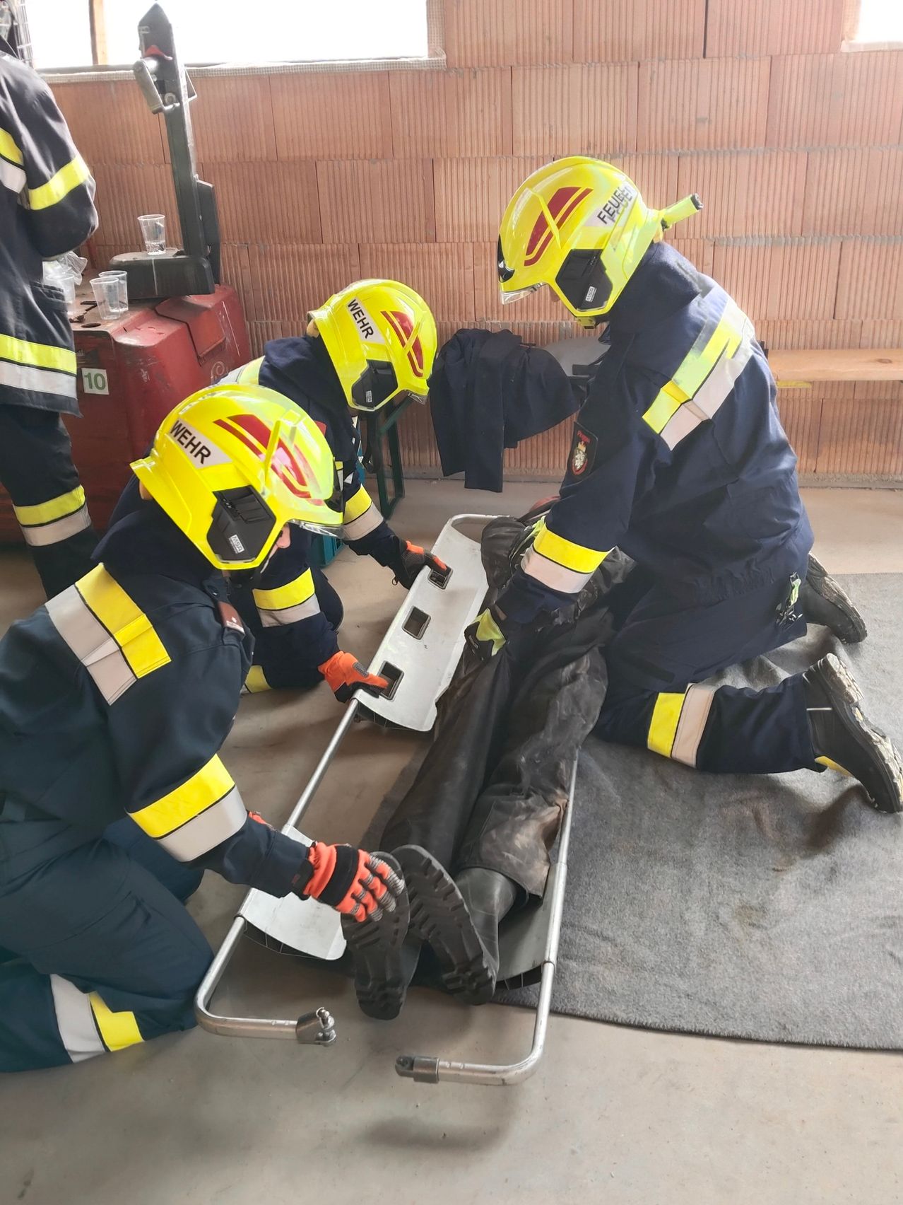 Drei Feuerwehrleute in gelben und blauen Uniformen retten eine Person auf einer Trage. Sie tragen Helme und Handschuhe, und einer von ihnen kniet auf dem Boden.