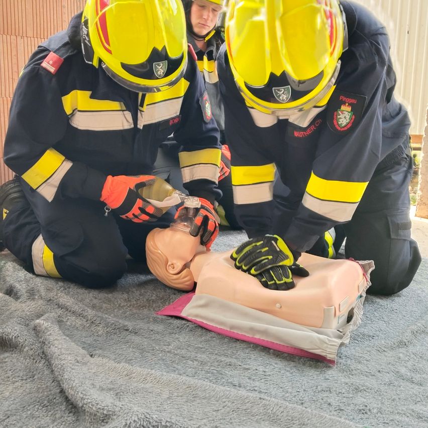 Zwei Feuerwehrleute üben CPR an einem Mannequin. Einer führt Brustkompressionen durch, während der andere Rettungsatmungen durchführt. Sie tragen gelbe Helme und schwarze Uniformen mit gelben Streifen.