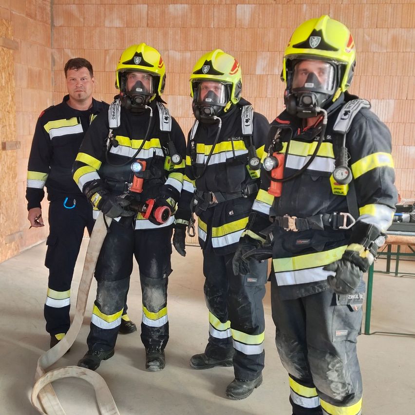 Vier Feuerwehrleute in voller Ausrüstung stehen in einem Raum, einer hält eine Feuerwehrschläuche. Dahinter ist eine Holzwand und eine Bank zu sehen.