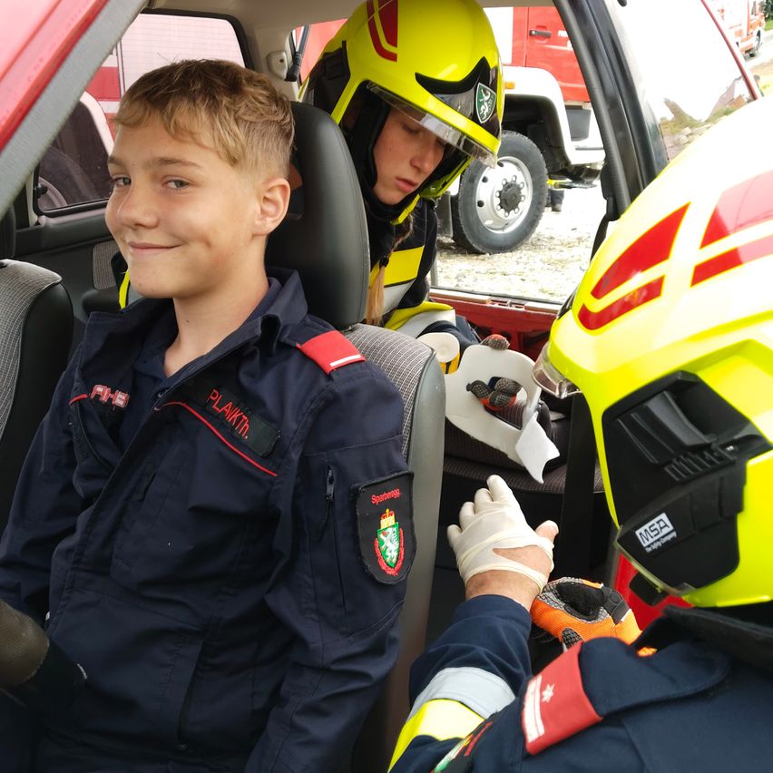 Ein lächelnder Junge in einer blauen Uniform sitzt in einem Fahrzeug, möglicherweise einem Feuerwehrwagen, mit einer weiteren Person in gelbem Helm in der Nähe.