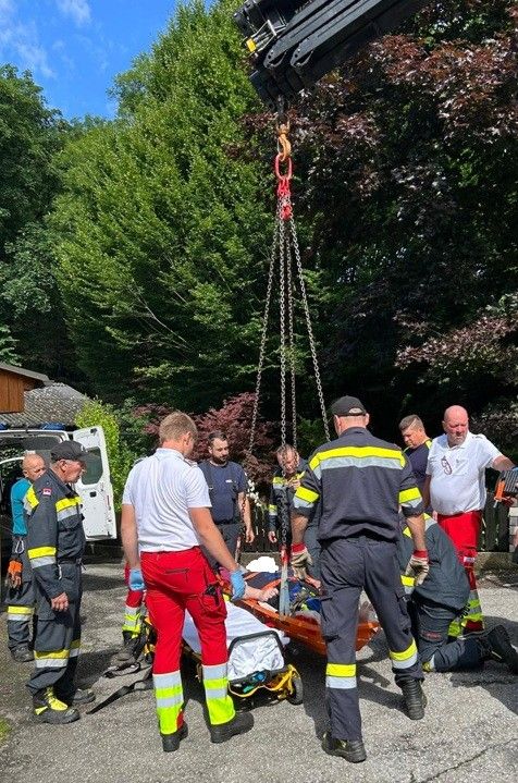 Eine Gruppe von Rettungskräften benutzt einen Kran, um eine Trage aus einem Baum zu senken. Die Szene spielt sich an einem Außenbereich ab, mit einem Van, Bäumen und einem Gebäude im Hintergrund.