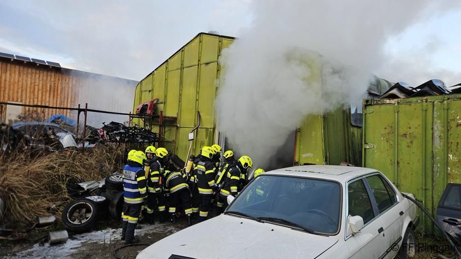 Eine Gruppe von Feuerwehrleuten arbeitet in der Nähe eines Containers, aus dem Rauch austritt. Sie befinden sich in der Nähe eines weißen Autos.