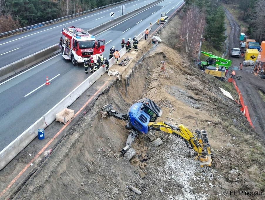 Eine Autobahn mit einer großen Ausgrabung, einem blauen Bagger und einem gelben Bagger an der Seite, mehreren Personen in Sicherheitsausrüstung, einem Feuerwehrwagen und anderen Fahrzeugen.