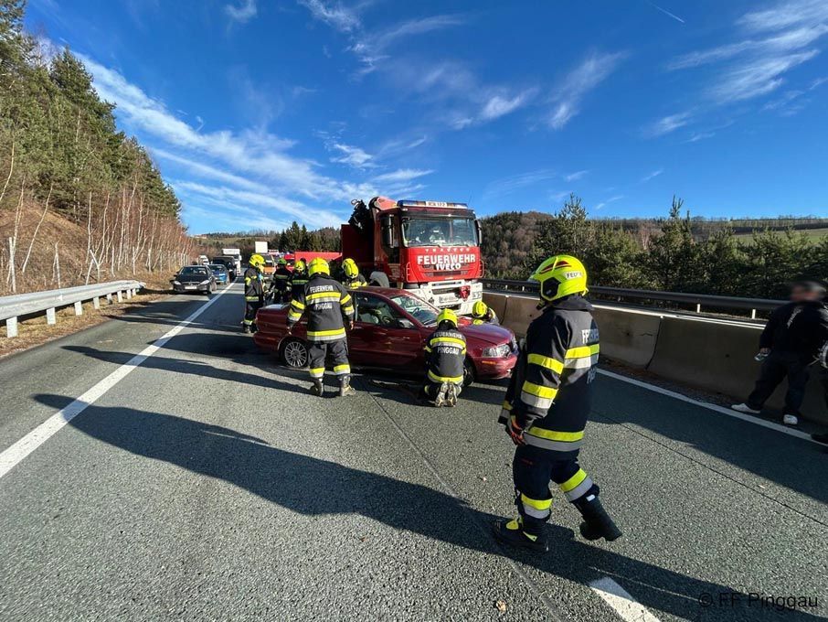 Feuerwehrleute in voller Ausrüstung kümmern sich auf einer Autobahn um einen roten Wagen. Sie arbeiten mit einem Feuerwehrwagen im Hintergrund an dem Auto.