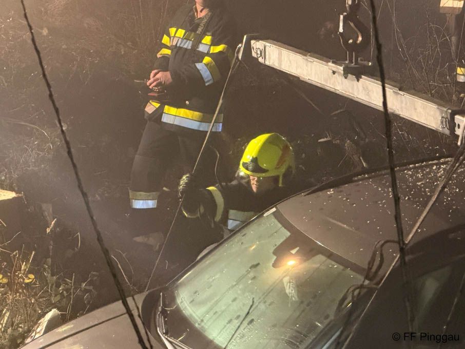 Zwei Feuerwehrleute arbeiten an einem beschädigten Auto, einer trägt einen gelben Helm. Sie verwenden einen Haken, um das Fahrzeug anzuheben.